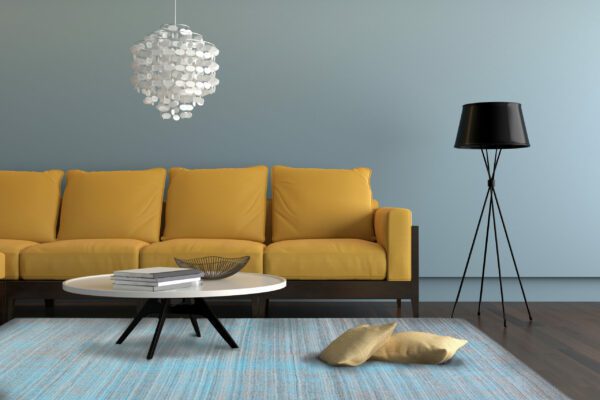 Close up view of a living room showing a yellow orange sofa, with a lamp, ornate pendant and a table with decoration. There is a gray carpet with two pillows on it. Light blue wall surrounding the scene with wooden floor. Horizontal composition.