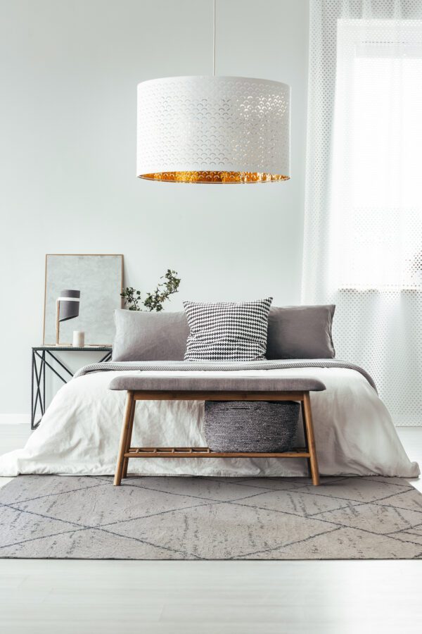 White lamp above wooden bench on carpet in designer bedroom with window and pillows on bed